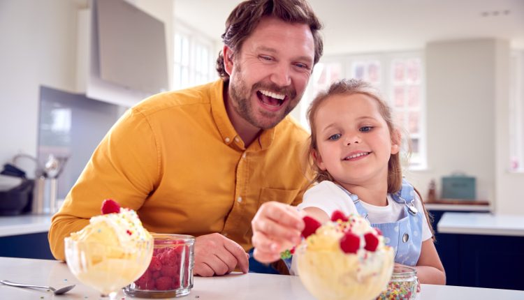 Father And Daughter In Kitchen Decorating Ice Cream Dessert With Cream And Raspberries